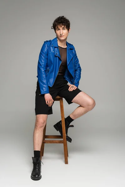 Comprimento total da pessoa pansexual em jaqueta de couro azul e botas pretas sentadas em banquinho de madeira em fundo cinza — Fotografia de Stock
