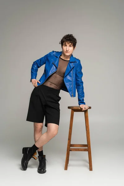 Voller Körperlänge bigender Person in blauer Lederjacke und schwarzer Shorts, die in der Nähe eines Holzhockers auf grauem Hintergrund posiert — Stockfoto