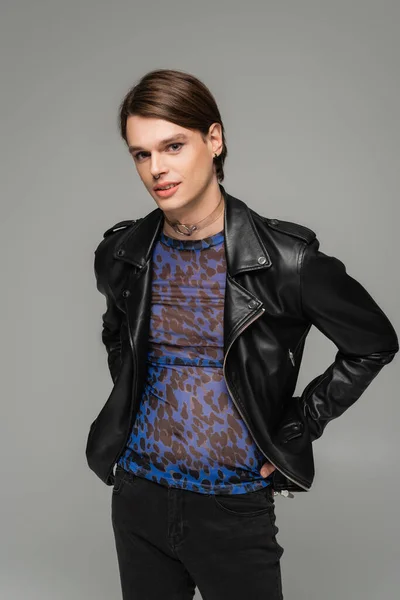 Junge nichtbinäre Person in Animal-Print-Top und schwarzer Lederjacke stehend mit den Händen an den Hüften isoliert auf grau — Stockfoto