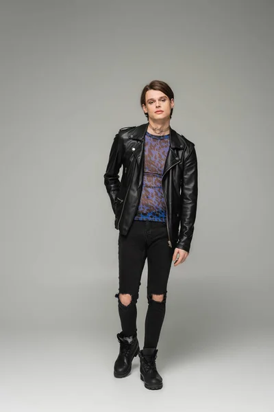 Full length of stylish nonbinary person in black leather jacket and ripped pants on grey background — Stock Photo