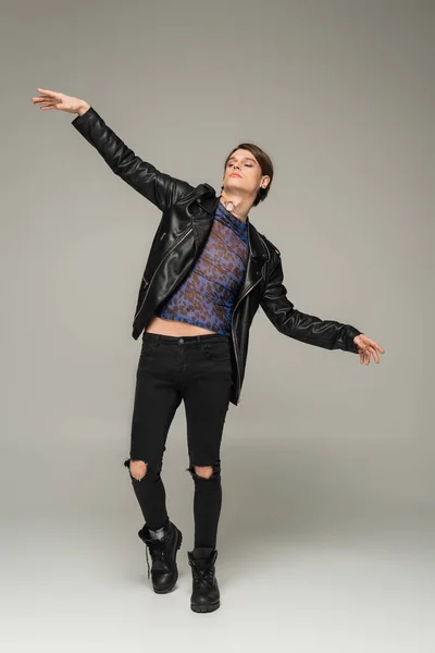 Full length of fashionable pansexual person in ripped pants and black leather jacket posing with outstretched hands on grey background — Stock Photo