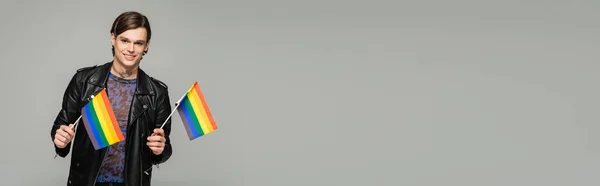Modelo pansexual alegre em jaqueta de couro preto segurando pequenas bandeiras lgbt isolado em cinza, banner — Fotografia de Stock