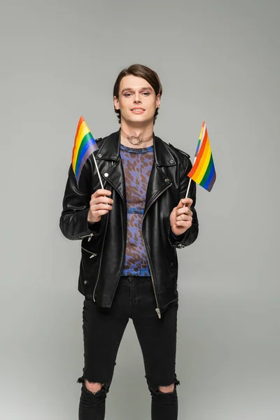 Cheerful and trendy pangender model in animal print top and black leather jacket holding small lgbt flags isolated on grey — Stock Photo