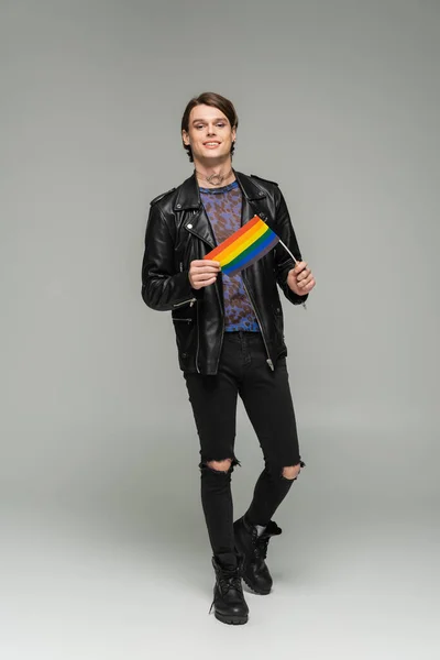 Full length of carefree pangender person in black ripped pants and leather jacket standing with small lgbt flag on grey background — Stock Photo