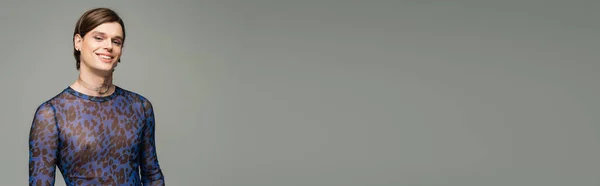 Giovane e spensierato bigender in top stampa animalier sorridente alla fotocamera isolata su grigio, banner — Foto stock