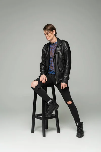 Full length of bigender model in black leather jacket and ripped pants posing near high stool on grey background — Stock Photo