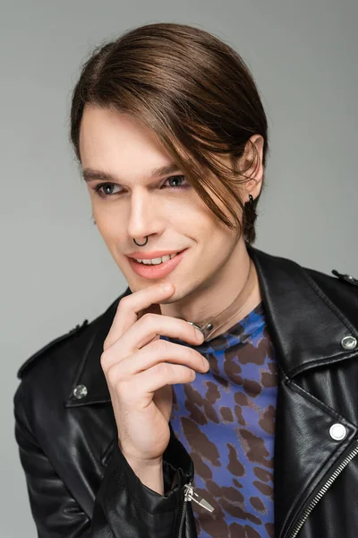 Portrait of smiling bigender model with makeup and nose piercing wearing black leather jacket isolated on grey — Stock Photo