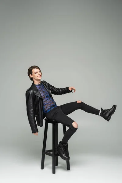 Carefree bigender person in black ripped pants and leather jacket posing on high stool on grey background — Stock Photo