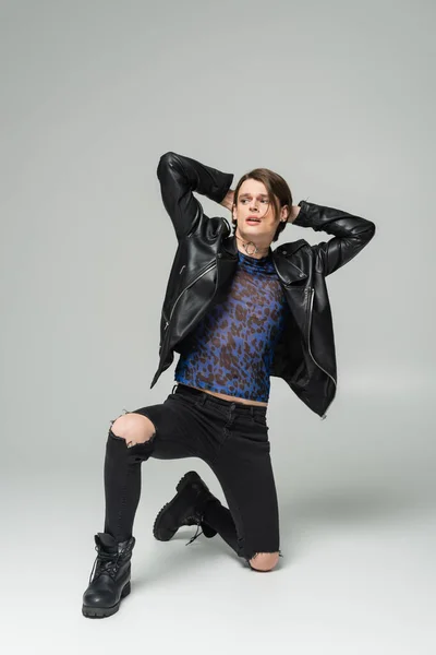 In voller Länge trendiges bigender Model in schwarzer Lederjacke und zerrissener Hose posiert mit den Händen hinter dem Kopf auf grauem Hintergrund — Stockfoto
