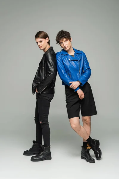 In voller Länge junge bigeschlechtliche Partner in blauen und schwarzen Lederjacken posieren auf grauem Hintergrund — Stockfoto