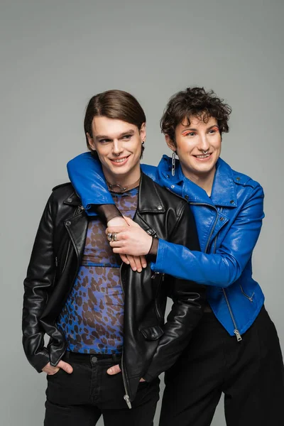Glückliche bigeschlechtliche Person in blauer Lederjacke umarmt stilvollen Partner isoliert auf grau — Stockfoto