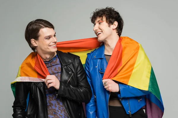 Alegres socios grandes en chaquetas de cuero posando con la bandera del arco iris aislado en gris - foto de stock