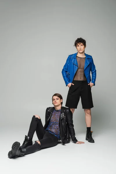 Giovane non binario in giacca di pelle blu in piedi con le mani in tasche vicino partner alla moda seduto su sfondo grigio — Foto stock