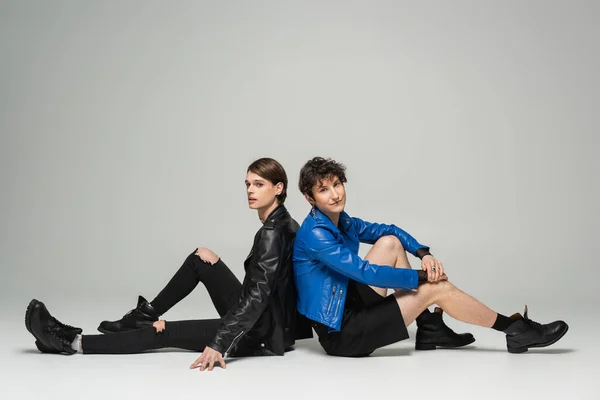 Fashionable pansexual partners in leather jackets sitting back to back on grey background — Stock Photo