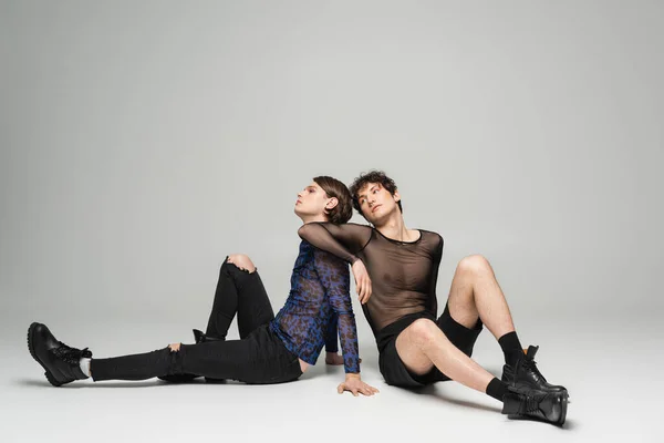 Full length of bigender partners in black leather boots and trendy tops sitting on grey background — Stock Photo