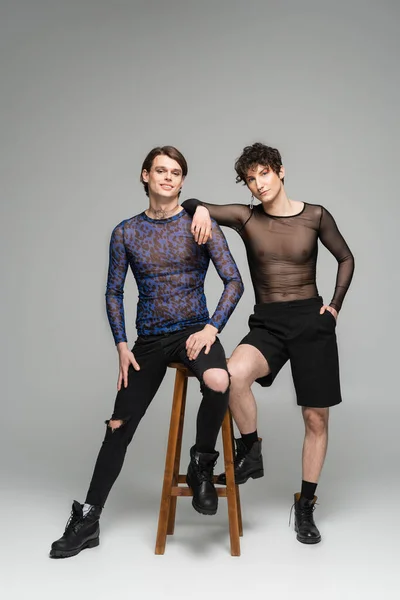 Full length of young and fashionable nonbinary models posing near high stool on grey background — Stock Photo