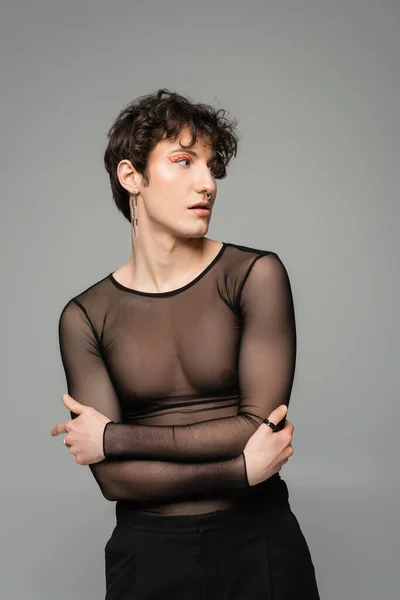 Young nonbinary person in makeup and black transparent top standing with crossed arms and looking away isolated on grey — Stock Photo