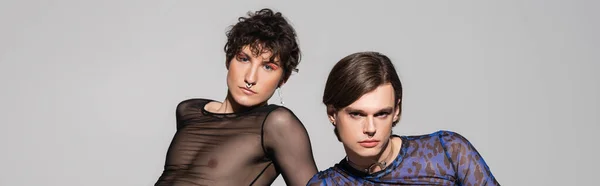 Junges pansexuelles Paar in modischer Kleidung und Make-up mit Blick auf Kamera isoliert auf grau, Banner — Stockfoto