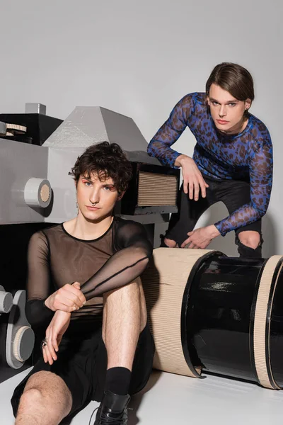 Junges Pangender-Paar in modischen Oberteilen posiert in der Nähe des Fotomodells auf grauem Hintergrund — Stockfoto