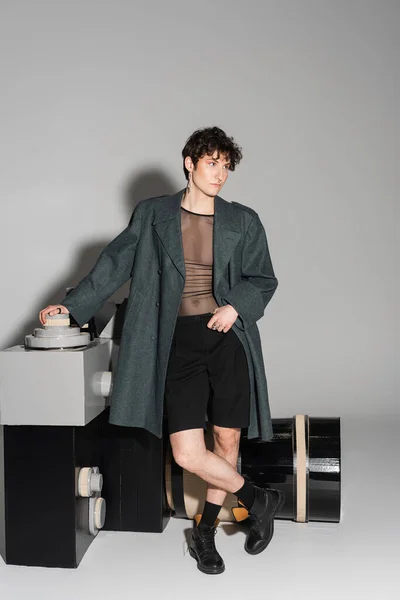 Full length of stylish nonbinary person in coat and black leather boots looking away near model of photo camera on grey background — Stock Photo