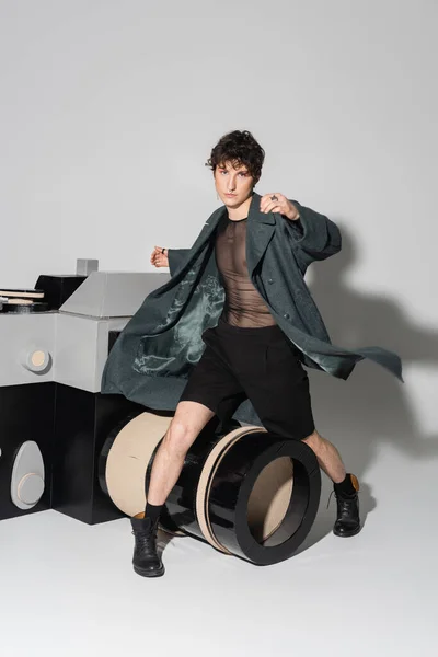 Full length of pansexual person in coat and black leather boots posing near huge model of photo camera on grey background — Stock Photo
