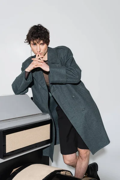 Persona pangender premuroso in posa cappotto con le mani serrate vicino modello di macchina fotografica su sfondo grigio — Foto stock
