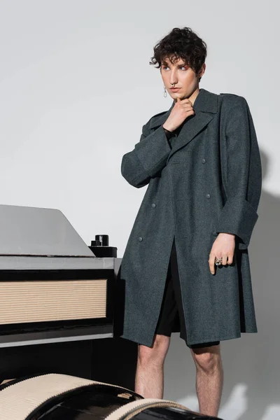 Stylish and thoughtful nonbinary person in coat looking away near model of photo camera on grey background — Stock Photo