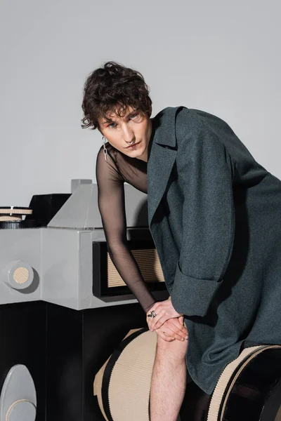 Trendige pansexuelle Person im Mantel posiert in der Nähe von riesigen Modell der Fotokamera isoliert auf grau — Stockfoto