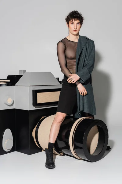 Fashionable nonbinary person in black transparent top and coat standing near huge model of photo camera on grey background — Stock Photo