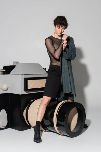 Young pansexual person in black shorts and coat standing near model of photo camera on grey background — Stock Photo