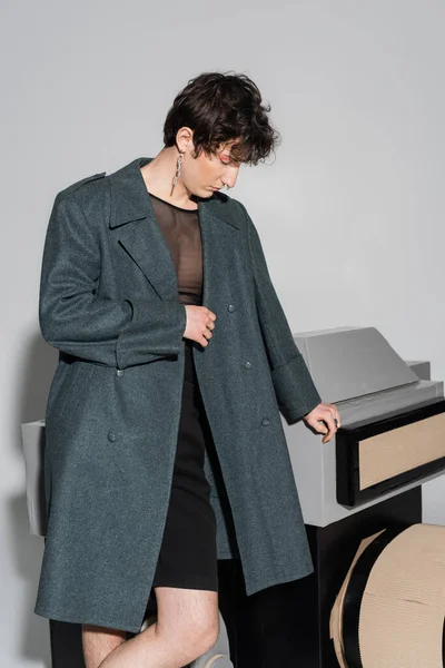 Junge und trendige pansexuelle Person im Mantel mit Blick auf ein riesiges Fotomodell auf grauem Hintergrund — Stockfoto