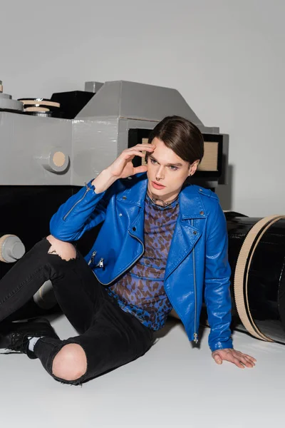 Dreamy pansexual person in blue leather jacket sitting near huge model of photo camera on grey background — Stock Photo
