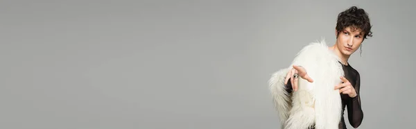 Brünette zweigeschlechtliche Person in weißer Kunstpelzjacke, die wegschaut und mit Fingern auf grau isoliert zeigt, Banner — Stockfoto