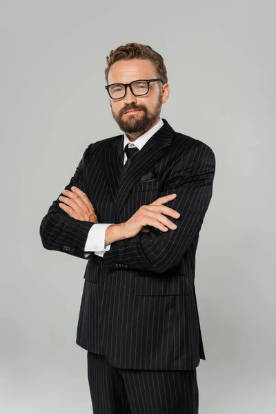 Веселий бізнесмен у формальному одязі та окулярах позує схрещеними руками ізольовано на сірому — стокове фото