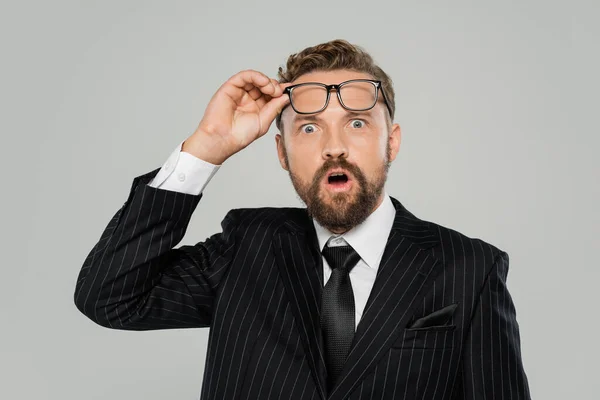 Шокований бізнесмен у формальному носінні регулює окуляри і дивиться на камеру ізольовано на сірому — стокове фото