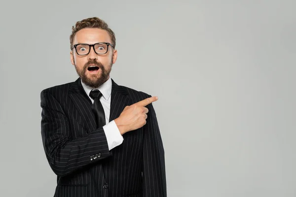 Astonished businessman in formal wear and glasses pointing away isolated on grey — Stock Photo