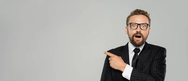 Изумленный бизнесмен в формальной одежде и очках, указывающих на изолированные на сером, баннер — стоковое фото