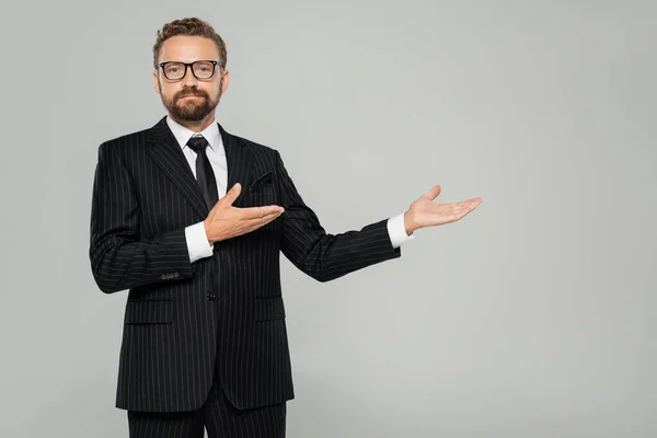 Бородатый бизнесмен в формальной одежде и в очках, указывающих на руки, изолированные на сером — стоковое фото