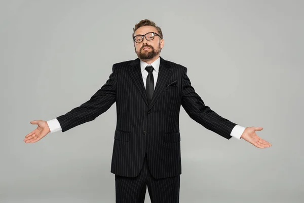 Homme d'affaires confus en tenue formelle et lunettes regardant la caméra tout en montrant un geste d'haussement d'épaules isolé sur gris — Photo de stock