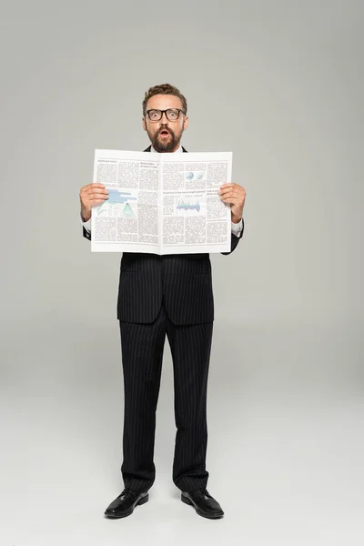 Full length of astonished businessman in suit and glasses holding newspaper on grey — Stock Photo