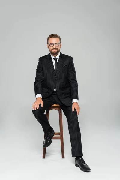 Full length of happy businessman in suit and glasses sitting on wooden high chair on grey — Stock Photo