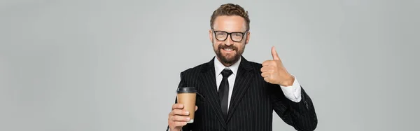 Happy businessman in glasses and suit holding paper cup and showing thumb up isolated on grey, banner — Stock Photo