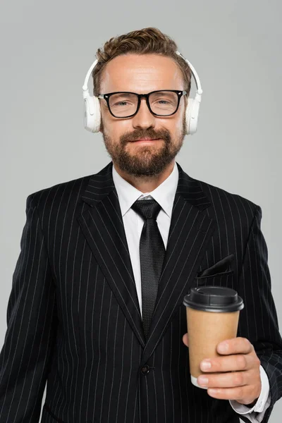 Homem de negócios feliz em óculos e fones de ouvido segurando copo de papel isolado em cinza — Fotografia de Stock