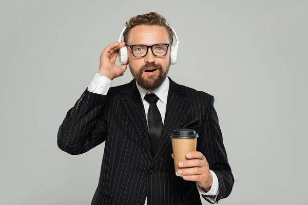 Изумленный бизнесмен в очках и костюме, держащий бумажную чашку, слушая музыку, изолированную на сером — стоковое фото