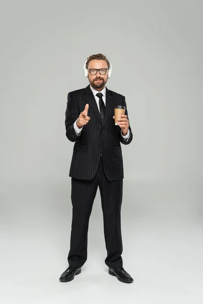 In voller Länge glücklicher Geschäftsmann in Brille und Anzug, Pappbecher haltend und mit dem Finger auf grau zeigend — Stock Photo