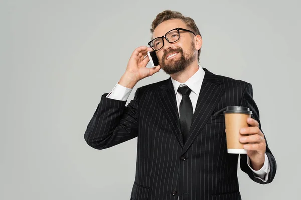 Homme d'affaires heureux en lunettes et costume parler sur smartphone et tenant tasse en papier isolé sur gris — Photo de stock