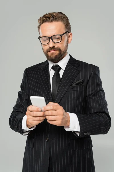 Bärtiger Geschäftsmann mit Brille und Anzug tippt auf Smartphone isoliert auf grau — Stockfoto