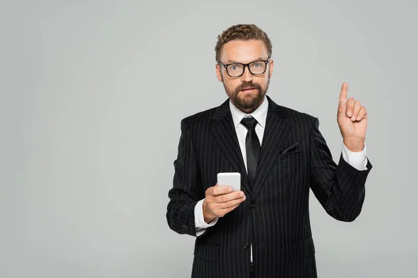 Hombre de negocios barbudo en gafas y traje sosteniendo teléfono inteligente y apuntando con el dedo aislado en gris - foto de stock