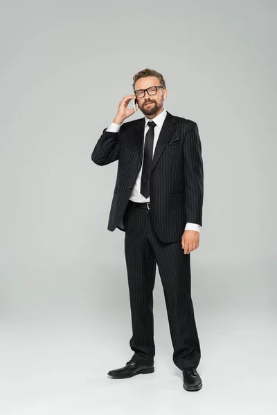 Full length of businessman in suit and glasses talking on smartphone on grey — Stock Photo