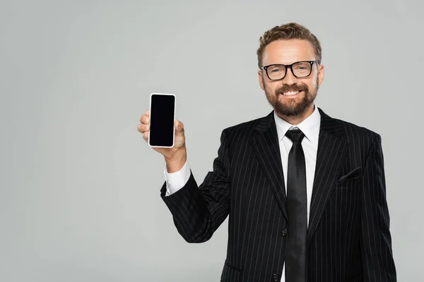 Homme d'affaires heureux en costume et lunettes montrant smartphone avec écran blanc isolé sur gris — Photo de stock